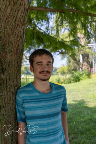 Relaxed Portrait in the Shade Young man in a striped blue shirt leaning against a tree in dappled afternoon shade
