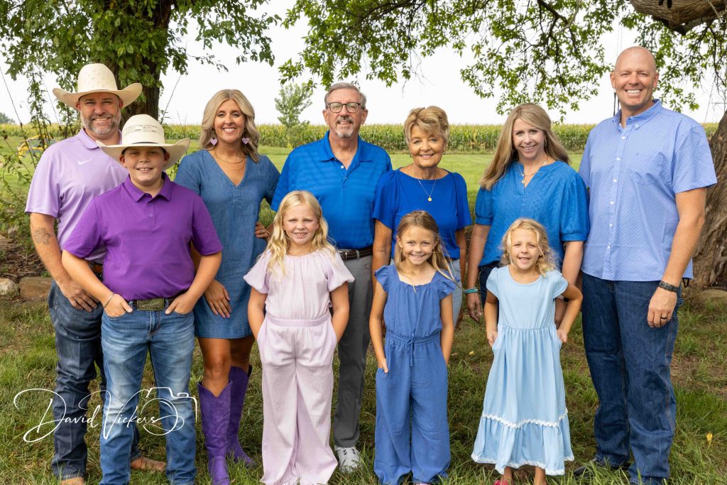 Family Photographer in Olathe, KS Three generations of a family in coordinated blue and purple standing together under a shade tree beside a Kansas cornfield