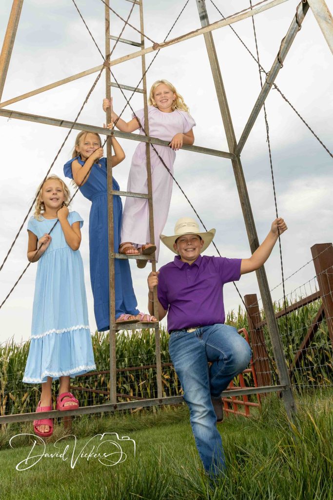 Family Photographer in Olathe, KS Four cousins in blue and lavender climbing an old steel windmill tower at the edge of a cornfield