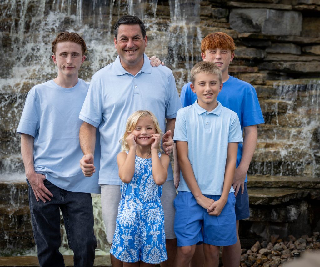 Family Photographer in Overland Park, KS Father with his four children in coordinated blue in front of a cascading stone waterfall