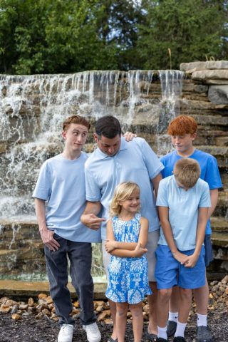 Family Portrait at the Falls Family of five gathered in front of a stone waterfall surrounded by summer greenery