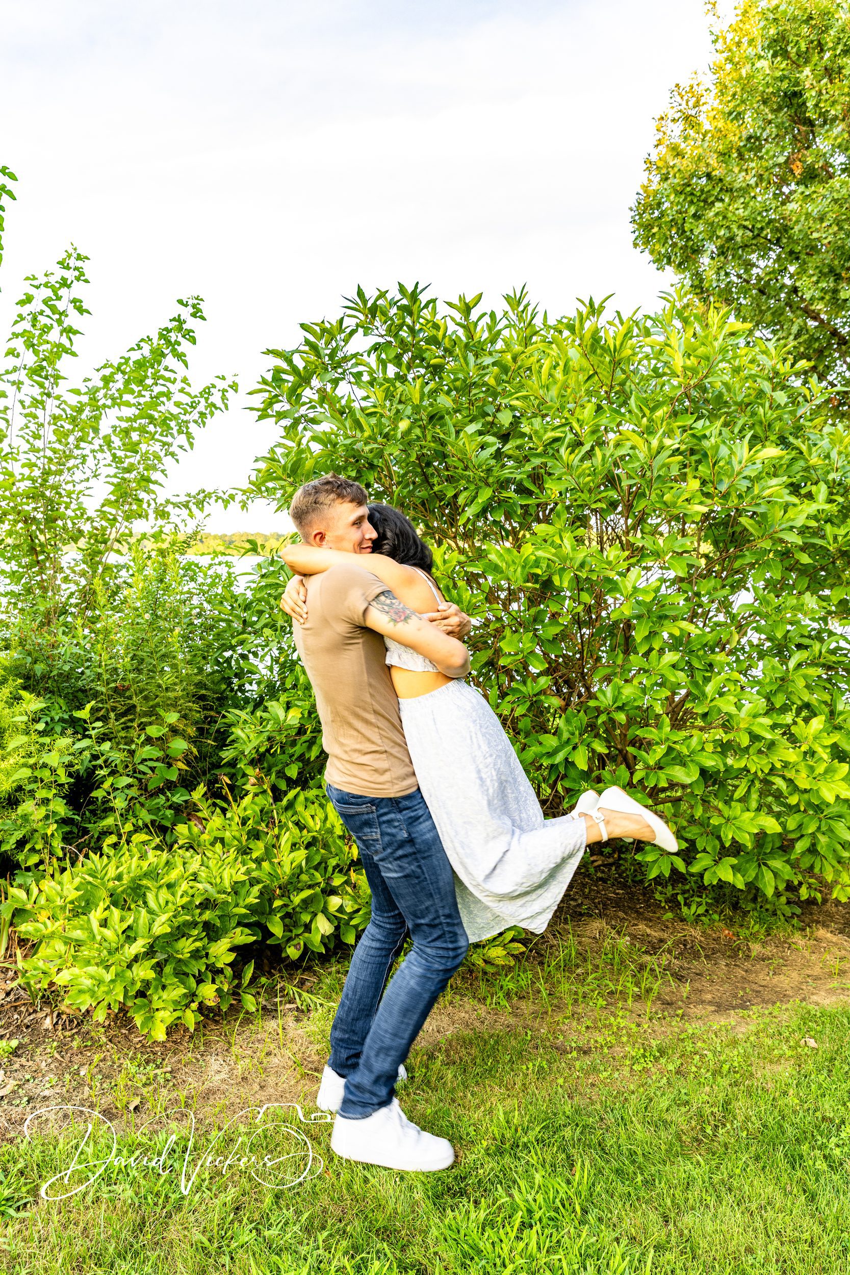 Lifestyle Family Photography in Kansas City: What to Expect A man lifting his fiancee off the ground in a joyful hug beside a lake
