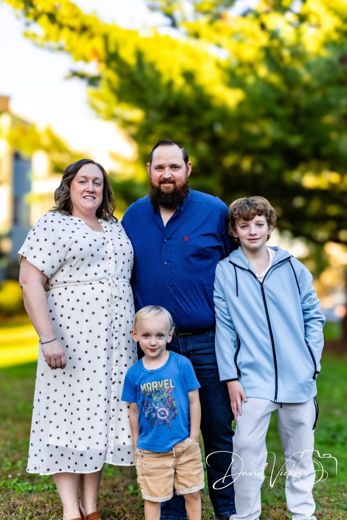 Mom, dad and their two sons standing close together on a lawn under pine trees in warm evening light